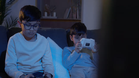 Two-Young-Boys-Sitting-On-Sofa-At-Home-Playing-With-Computer-Games-Console-On-TV-Holding-Controllers-Late-At-Night-1